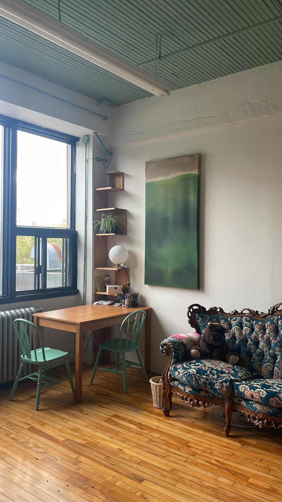 a bright corner of the studio next to a big window. you can see one end of an antique blue floral couch with a stuffed hippo, an art table with two chairs under the window, a large deep green & sand painting, and a set of shelves in the corner with art supplies