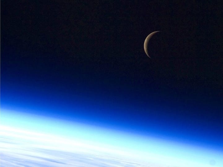 sliver of a waning crescent moon in a deep blue starless sky while the earth's atmosphere glows bright white below