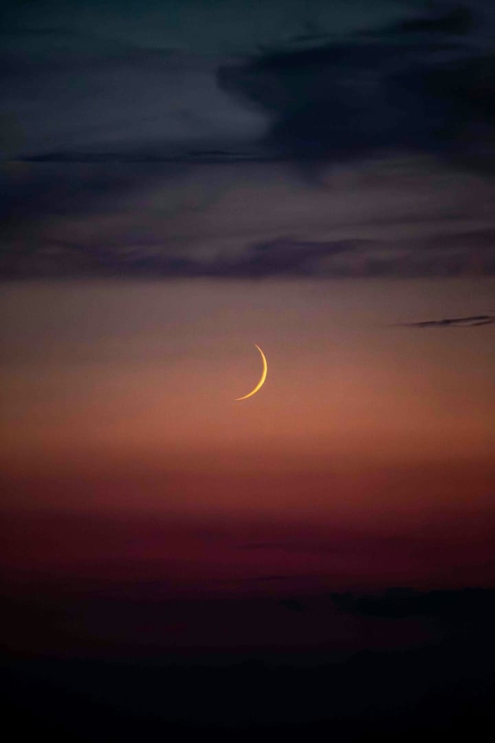yellow sliver of a waxing crescent moon over sunset colours of peach, fuschia and deep purple